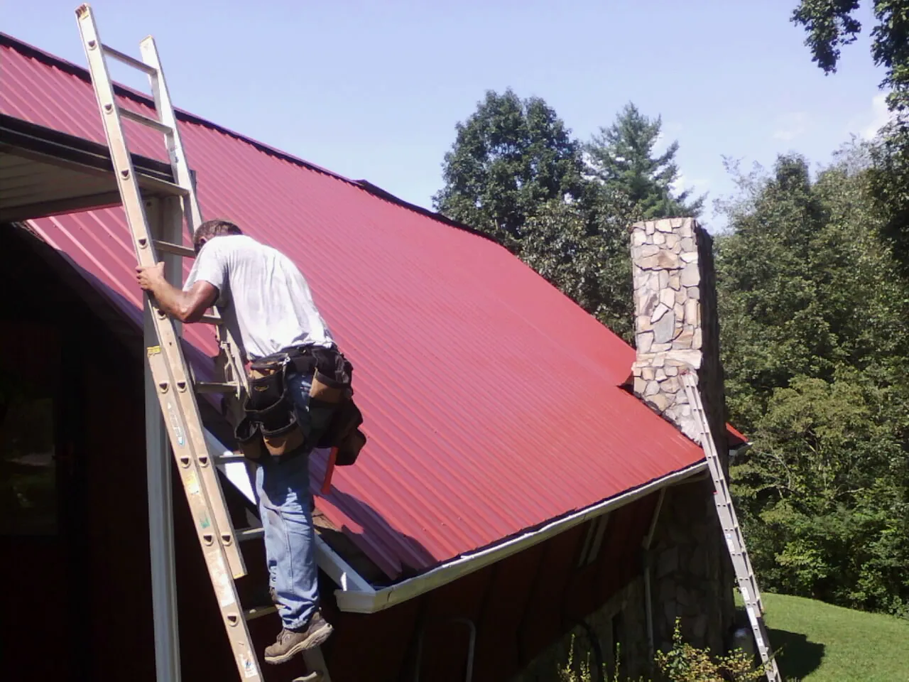Roofer on a ladder beside a red metal roof and stone chimney — Soffit & Fascia Repair by Roofing Brothers in Lake George
